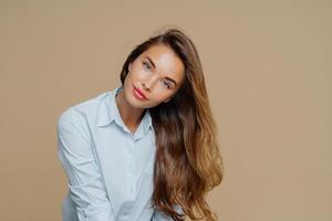 Portrait of pleasant looking female tilts head on right, has long well cared hair, wears blue shirt, has appealing appearance, makeup, isolated over brown background with blank space for information photo