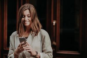 Horizontal shot of pretty cute brunette female model shares multimedia files via cell phone, dressed in elegant clothes, reads positive news in internet website, connected to wireless internet. photo