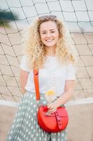 Vertical shot of good looking curly female wears t shirt and skirt, has small bag, holds cocktail with ice, stands near tennis net on beach, has good rest. Lovely woman with appealing charming smile photo
