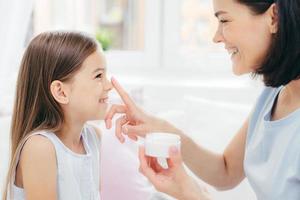 Close up shot of pretty cheerful female recieves beauty procedure from affectionate mother. Attractive young woman applies beauty cream on daughters nose. Motherhood and upbringing concept.