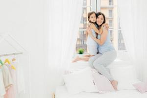 Indoor shot of pleasant looking female mother embraces her small kid, sit on window sill near bed, enjoy togetherness, have friendly relationship, pose in spacious light bedroom. Motherhood concept photo