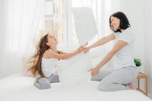 Parenthood and family concept. Attractive female mother with positive smile, spends free time with her daughter, have pillow fight, pose against bedroom interior, enjoys leisure time during weekend photo