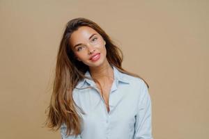 Portrait of pleasant looking cheerful female has long hair, tilts head and looks with smile at camera, wears blue shirt, uses cosmetics for makeup, poses against brown background, blank space aside photo