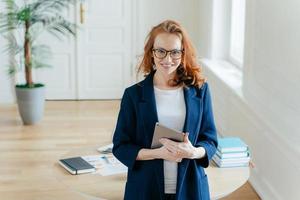 Horizontal shot of proud CEO checks email on digital touchpad, looks at camera with satisfied expression, wears spectacles, stands in modern office, enjoys online communication on modern device photo