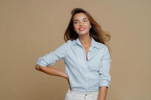 Shot of pretty cheerful European woman keeps one hand on waist, looks aside, has charming smile wears shirt and trousers, has slim perfect figure, enjoys shopping day, isolated over brown background photo