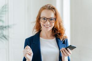 Photo of happy female leader with ginger hair, wears spectacles, updates app on modern cell phone, wears elegant clothes, poses in modern office, reads incoming message from business partner