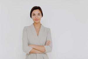 Prosperous successful business lady wears elegant wear, crosses hands while stands indoor at office over white wall, looks directly at camera. Female entrepreneur models indoor, waits for partner photo