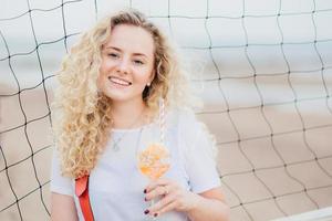 Optimistic young female model with curly light hair, dressed in casual white t shirt, drinks fresh cocktail, enjoys good rest at beach, stands near tennis net. People, beauty and rest concept photo