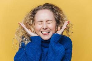 Happy joyful curly female with healthy skin, laughs loudly, keeps eyes shut, being in high spirit, hears funny jokes from interlocutor, wears blue sweater, poses against yellow studio background photo