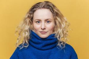 Indoor shot of good looking of serious blue eyed female wears blue warm sweater, looks confidently at camera, isolated over yellow background. Studio shot of beautiful curly woman models in studio photo
