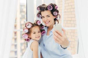 Attractive small kid with positive expression, charming smile stands near her mother, make selfie with modern mobile phone, pose against window background in room. Parenthood and beauty concept photo