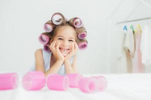 Portrait of cheerful female kid keeps hands under chin, has curlers on hair, going to have nice hairstyle, poses against white background, has charming smile, being in good mood. Children and beauty photo