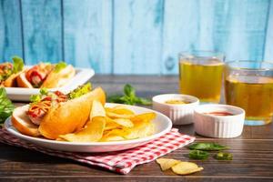 Delicious homemade hotdogs with dips sauces, chips and glasses of beer on wooden table. Top view. Copy space. photo