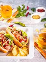 deliciosos perritos calientes caseros con salsas, patatas fritas y vasos de cerveza sobre una mesa de madera. vista superior. copie el espacio foto