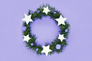 Christmas wreath made of fir-tree branches on very peri trend color background. Wreath decorated violet fluffy pom poms and white stars. Flat lay, top view, copy space. Color of new year 2022 concept.