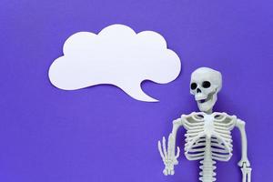 Skeleton on violet background with white blank paper cloud speech bubble. Anatomical plastic model human skeleton with talk text balloon. Empty dialog cloud, mockup, copy space. Purple Halloween. photo