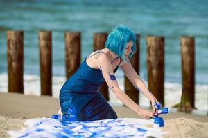 Artistic blue-haired woman performance artist smeared with gouache paints on large canvas on beach photo