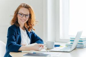 Self confident young female director plans work process, writes main theses in notepad, rewrites information from laptop computer, drinks tea, poses over office interior. Technology, occupation photo