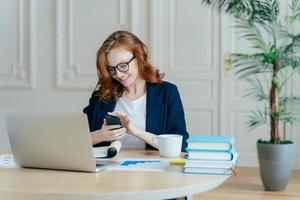 Technology and communication concept. Red haired young female checks email box on modern cell phone, has pleasant smile, studies documents, owns big corporation, uses laptop computer for work photo