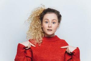 Pretty young female in bewilderment wears red warm loose sweater, indicates blank copy space of clothes for your advertising content, has curly pony tail, isolated over white studio background photo