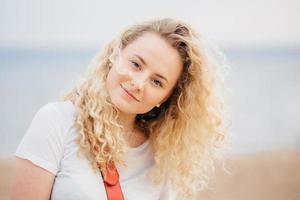 Outdoor shot of curly beautiful young female with fresh skin, looks positively at camera, wears casual white t shirt, poses against blurred seaside background. People and recreation concept. photo