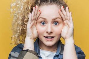 primer plano de una linda mujer emocional con piel sana y ojos azules, mantiene las manos en la cabeza, mira escrupulosamente y sorprendentemente a la cámara, nota algo, aislado sobre fondo amarillo foto