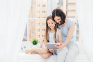 Portrait of pretty female embraces her little daughter, watch cartoon on digital tablet, connected to wireless internet at home, sit on window sill with white curtains at home. Children and parents photo