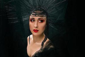 portrait of young beautiful woman like witch with red lips and black crown on dark background. Black dark beauty, Halloween concept photo