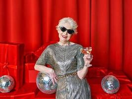 mujer elegante con estilo senior con una copa de vino espumoso sobre fondo rojo. fiesta, moda, celebración, concepto anti edad. foto