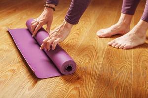 las manos de una mujer doblan una alfombra lila de yoga o fitness después de un entrenamiento en casa en la sala de estar. foto