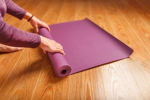 A woman's hands fold a lilac yoga or fitness mat after a workout at home in the living room. photo