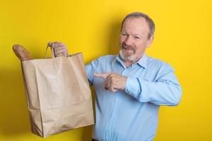 smiling handsome senior men holding a paper bag with purchases in his hand, pointing at it with the other hand photo