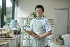 Portrait of an Asian male shopkeeper smiling and looking at camera, arranges natural products at refill store, zero-waste grocery, and plastic-free, eco environment-friendly, sustainable lifestyles. photo
