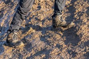 Piernas en pantalones grises y botas de montaña caminando hacia arriba en una colina fangosa a la luz del sol de la tarde foto