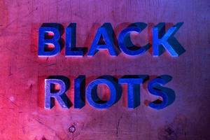 The words black riots laid with metal letters on flat plywood surface under red and blue police lights. Concept. photo