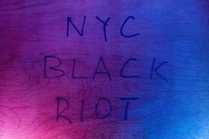 The words NYC black riot handwritten with black paint marker on flat plywood surface under red and blue police lights. Concept. photo