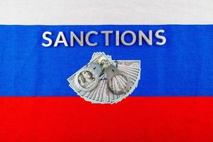 la palabra sanciones colocadas con letras de metal plateado en la bandera tricolor rusa cerca de los billetes en dólares y las esposas en una vista de ángulo alto foto