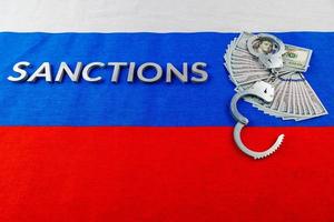 the word sanctions laid with silver metal letters on russian tricolor flag near dollar banknotes and handcuffs in linear perspective photo