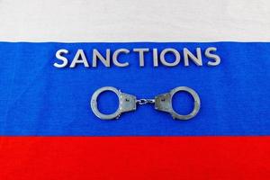 the word sanctions laid with silver metal letters on russian tricolor flag near handcuffs in directly above view photo