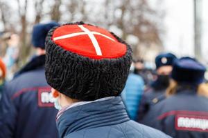 man in cossack hat with white cross on red watching blurry crowd of people photo