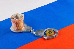 roll of russian paper rubles currency and bitcoin shiner chained with handcuffs on russian flag background photo