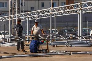 three Asian workers assembling metal construction by welding in front of car parking at sunny summer day under direct hard light photo