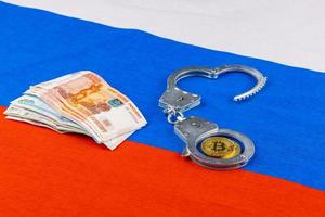 bunch of russian paper rubles currency and bitcoin shiner chained with handcuffs on russian flag background photo