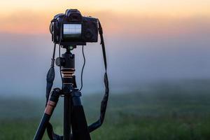 black digital camera on tripod shooting foggy morning landscape at summer with selective focus photo