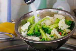 tap water flows over chopped cauliflower and broccoli in a colander above the sink while rinsing vegetables photo