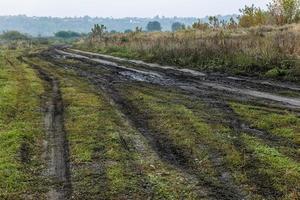 Mañana neblinosa otoñal paisaje rústico con camino de tierra en primer plano y pequeños edificios en la colina al fondo foto