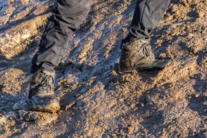 Piernas en pantalones grises y botas de montaña caminando hacia arriba en una colina fangosa a la luz del sol de la tarde foto