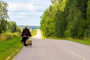 old woman with cart and black plastic bag walk away on side of rustic road at cloudy summer day photo