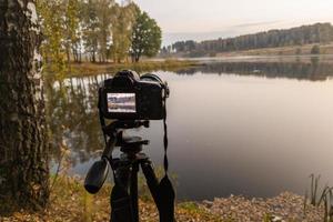 black digital camera on tripod shooting early foggy morning landscape at autumn lake with selective focus photo