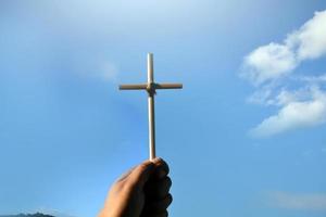 Small wooden cross holding in hand with cloudy and blue sky background, concept for love, hope, truth, faith, believe in Jesus, soft and selective focus.